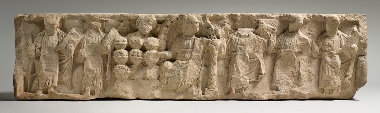 Long limestone relief fragment with worn standing figures and baskets of loaves at its center.