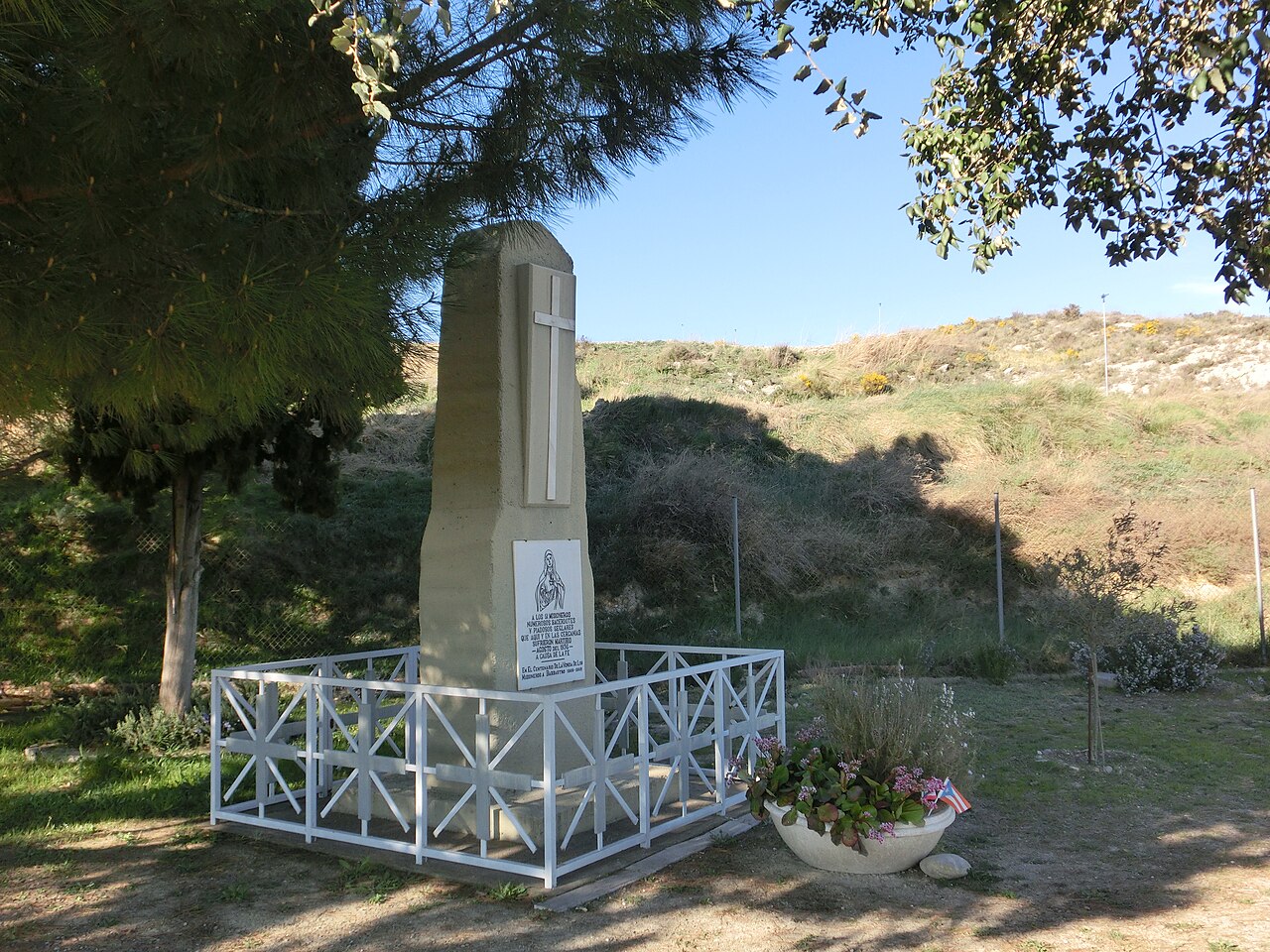 Roadside cross memorial near Barbastro, with trees and a public road behind it.